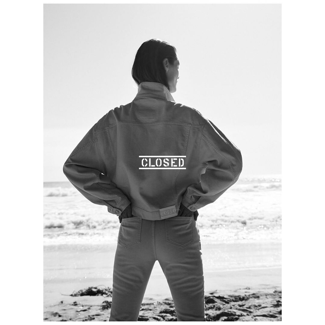 Back view of an adult woman in a denim jacket and jeans on a beach with the ocean and sky in the background in black and white.