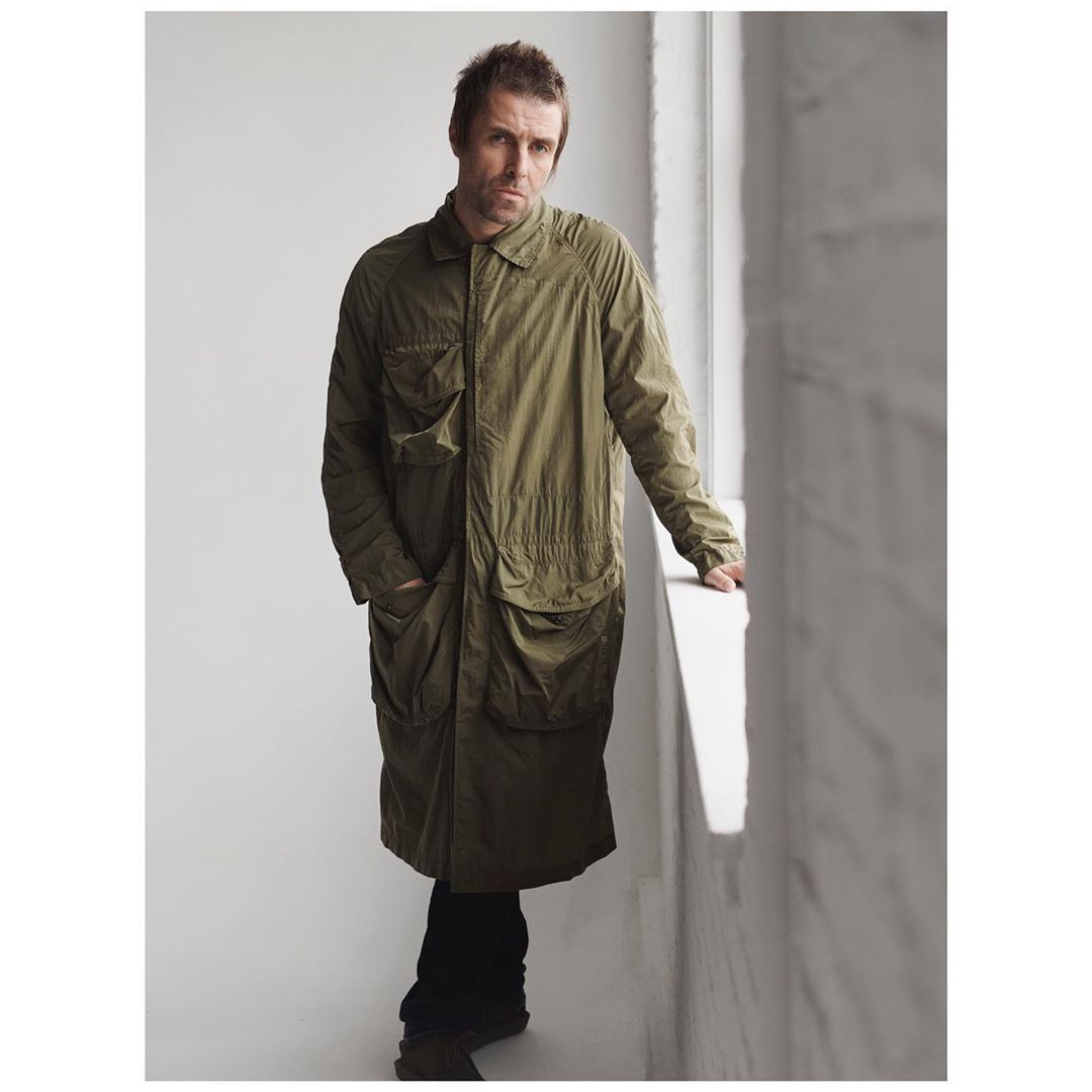 Adult male wearing a long green coat and black pants stands indoors near a window in a studio portrait with bright, natural light.