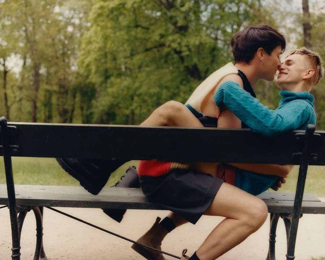 Two young men share a kiss while sitting on a park bench outdoors during the day surrounded by trees and green grass in summer.