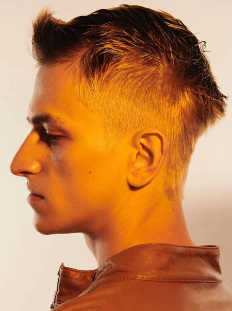 Close-up profile shot of a young adult man with short blond hair, wearing a brown leather jacket, side lit by warm light against a white background.