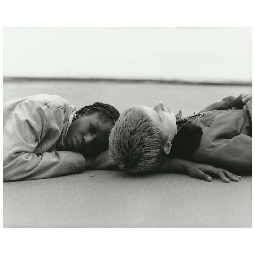 Monochrome photograph of two people lying head-to-head on a neutral surface, wearing workwear, in an outdoor setting, shot in a close-up.