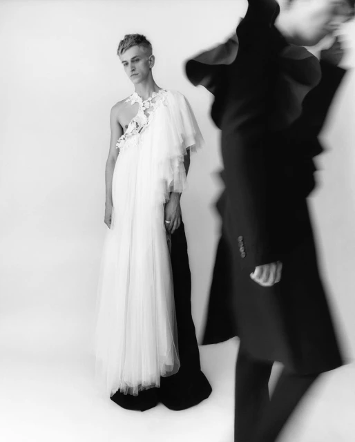 Monochrome fashion portrait of a young adult male wearing a white tulle gown with a floral detail alongside blurred figure in a black coat.