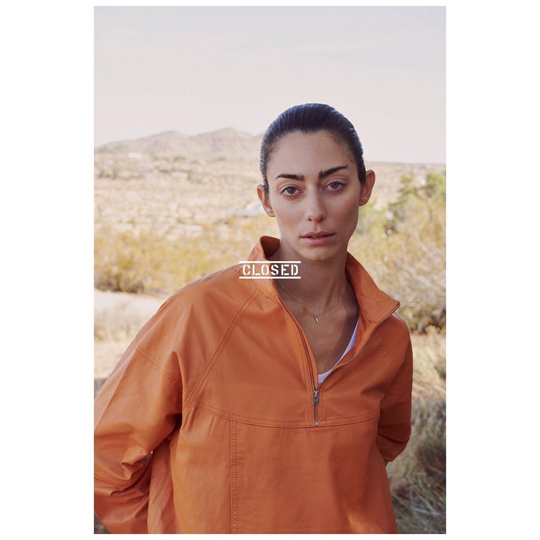 Portrait of a woman in an orange jacket against a desert backdrop, with fair skin, neutral expression, and slicked-back dark hair.