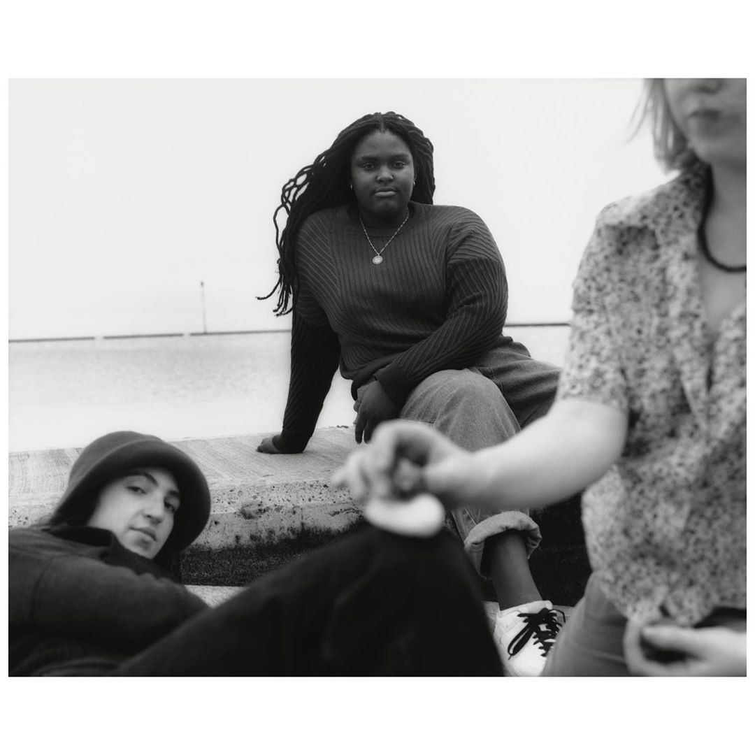 Monochrome portrait of three people outdoors on a concrete structure, featuring an African American woman with dreadlocks.