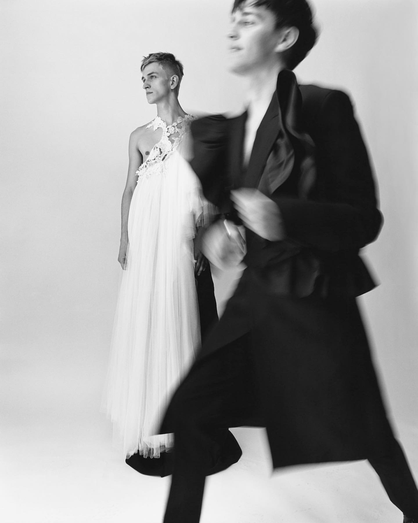 Two young adults in a high-contrast black and white studio shot. One wears a black suit, motion blur. The other wears a white gown.
