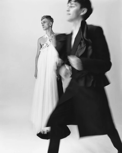 Two young adults in a high-contrast black and white studio shot. One wears a black suit, motion blur. The other wears a white gown.