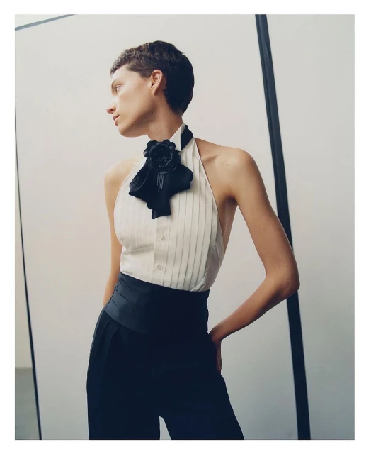 Fashion shot of a woman with short dark hair wearing a cream-colored, collared halter top with black high-waisted pants in a studio.