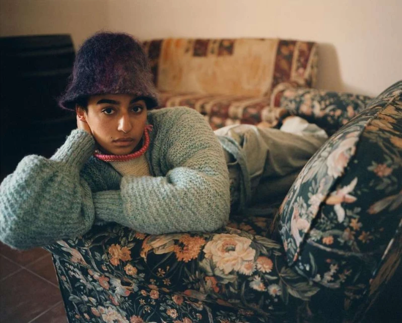 Young adult poses on a floral sofa wearing a light blue sweater, a red necklace, and a furry purple hat indoors with soft lighting.