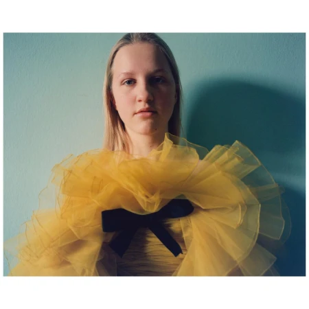 Portrait of a young woman wearing a yellow ruffled top with a black bow against a teal wall by Stefan Heinrichs