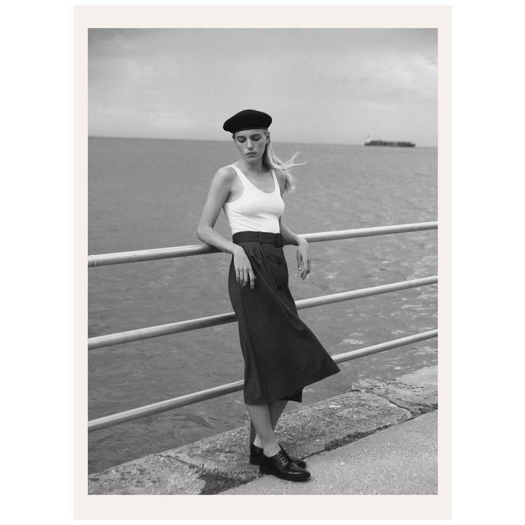 Woman in beret, tank top and skirt poses on pier in black and white near ocean. Fashion shot with sea and sky background.