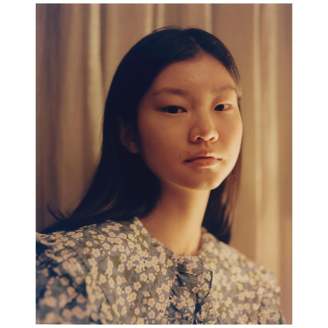 Portrait of an Asian woman with dark hair wearing a blue floral dress in a softly lit indoor setting with neutral-toned curtains.