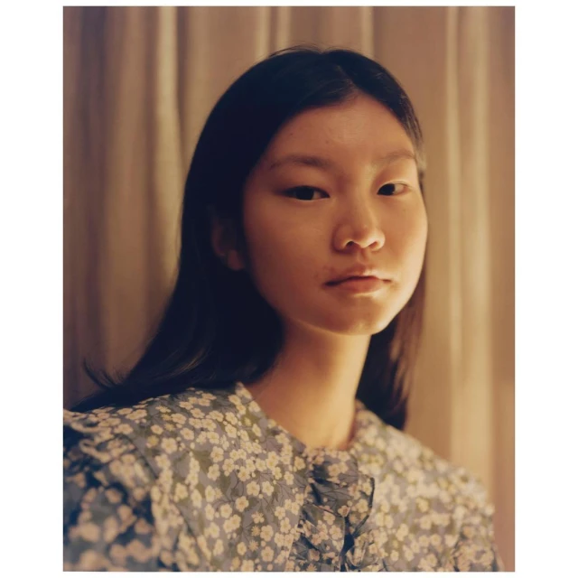 Portrait of an Asian woman with dark hair wearing a blue floral dress in a softly lit indoor setting with neutral-toned curtains.