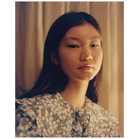 Portrait of an Asian woman with dark hair wearing a blue floral dress in a softly lit indoor setting with neutral-toned curtains.