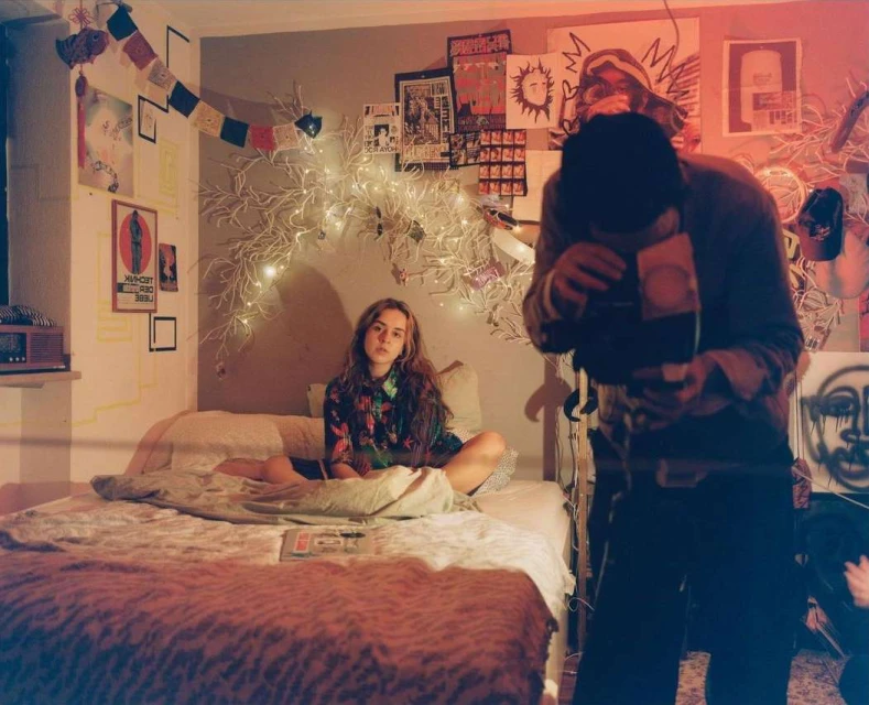 Two people inside a bedroom with posters on the wall, one seated on the bed and another person standing and holding what appears to be a camera.