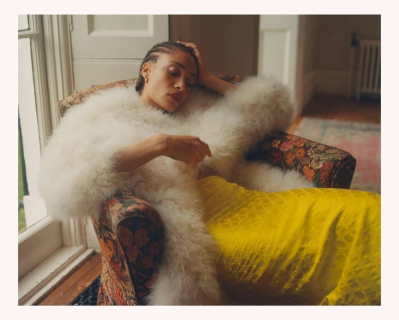 Woman with cornrows lounges in floral armchair wearing white feather coat and yellow dress, bathed in soft, natural window light.