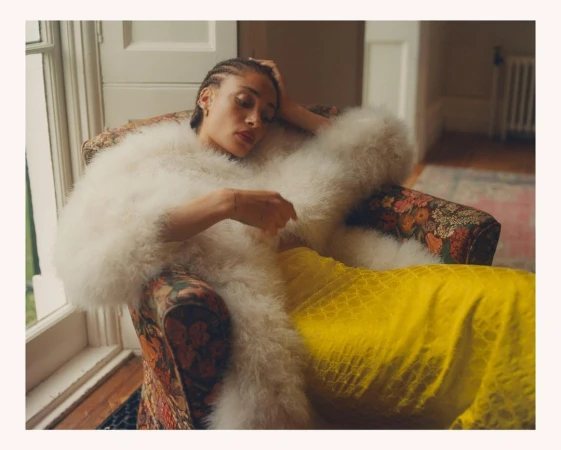 Woman with cornrows lounges in floral armchair wearing white feather coat and yellow dress, bathed in soft, natural window light.