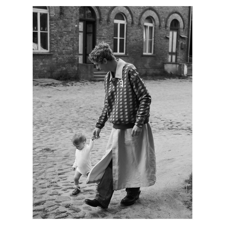 A black and white photo of a person in a patterned sweater and apron holding the hand of a young child on a cobblestone path.