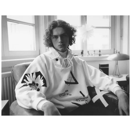 Young man with curly hair wearing a patterned white hoodie seated in a leather armchair indoors