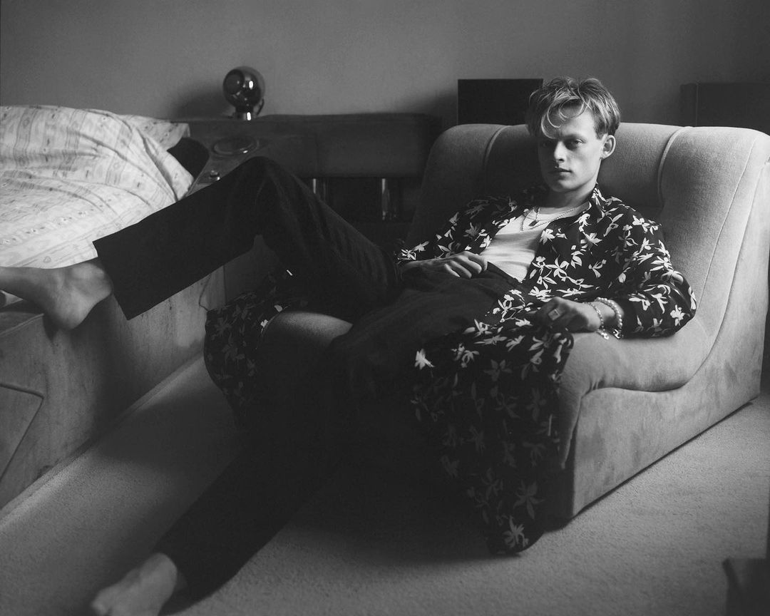 Black and white portrait of a young adult male lounging in an armchair wearing a floral print robe in an indoor setting.
