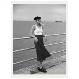 Monochrome portrait of a woman leaning on a metal railing by the sea, wearing a beret, tank top, skirt, and dark shoes.