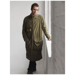 Liam Gallagher wearing a long green utility coat standing indoors by a bright window by photographer Stefan Heinrichs