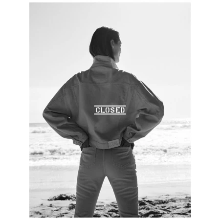 Model standing on a sandy beach wearing a denim jacket with text on the back, photographed from behind.