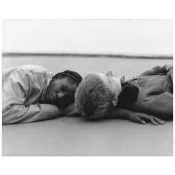 Black-and-white portrait of a young Black woman and a young white man lying on a sandy beach outdoors