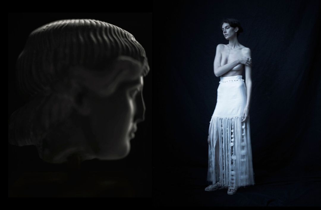 Monochromatic portrait of a classical bust alongside a woman in a long skirt, set against a stark black backdrop in a studio.