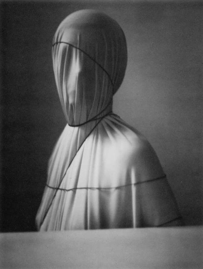 Monochrome portrait of a figure draped in fabric, head entirely covered, secured with dark string against a dark, soft-focus background.
