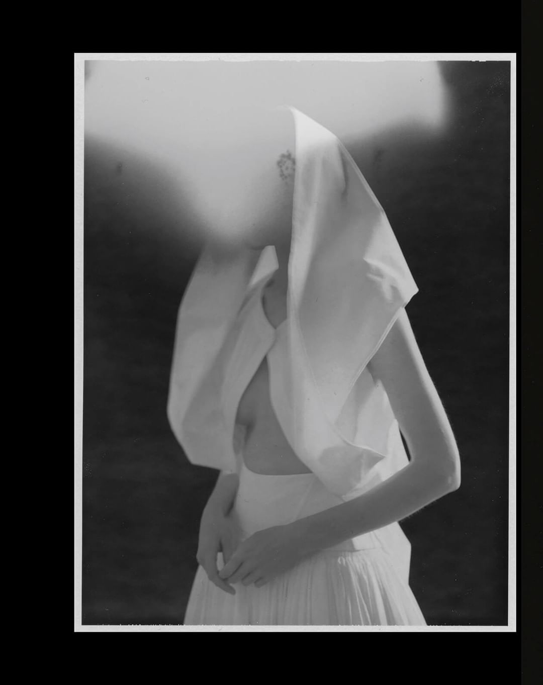 Black and white portrait of an adult female wearing a white gown with a hood, posing in a photo studio with a dark background.