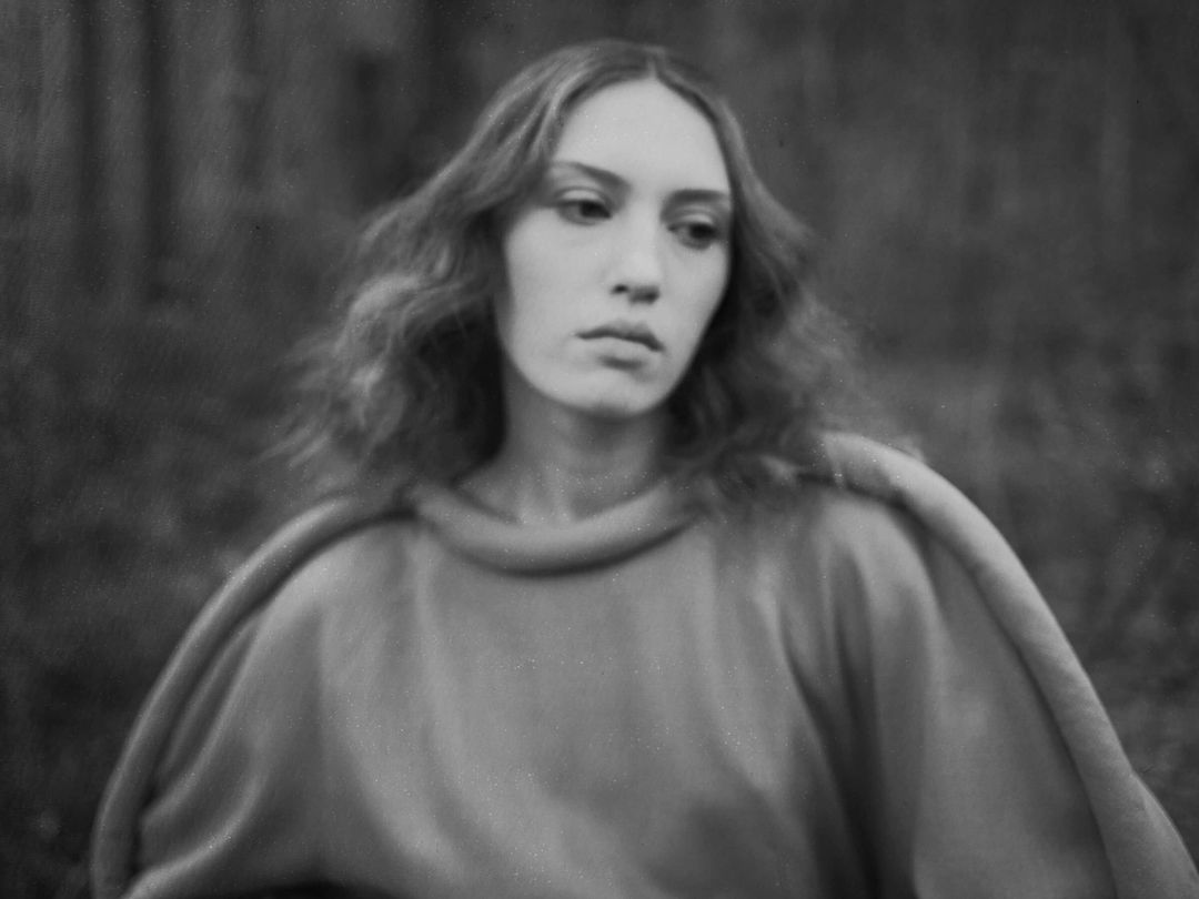 Black and white portrait of a young adult woman with long, wavy hair wearing a simple dress, set against a blurred nature background.
