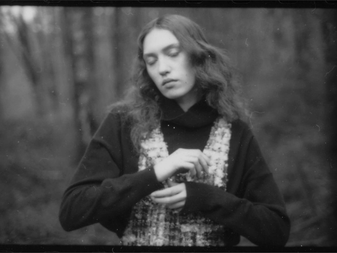 Monochrome portrait of a young adult female in a sweater and vest against a blurred forest background with a soft, desaturated aesthetic.