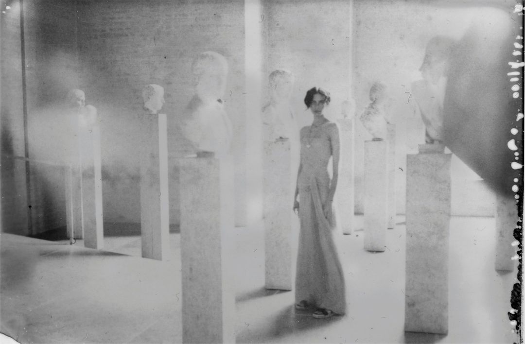 Monochromatic photo of a slender woman in a flowing gown standing among classical busts in an airy indoor space with muted tones.