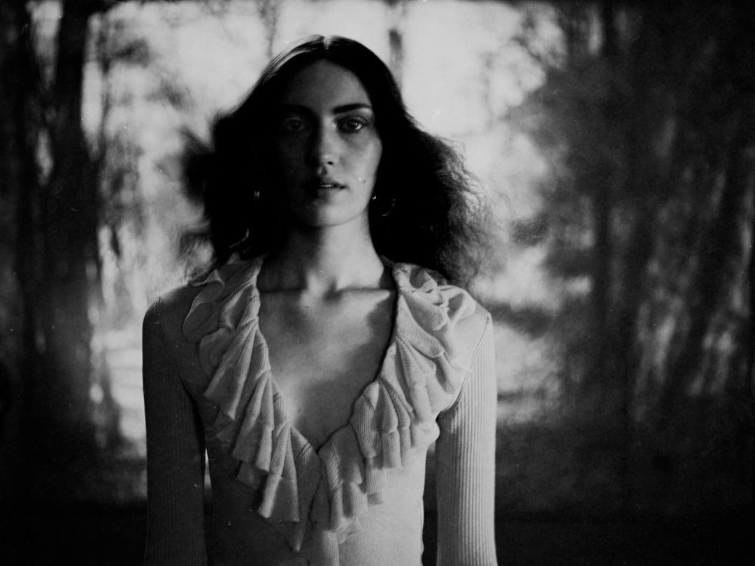 Monochrome portrait of an adult woman with long dark hair, wearing a ruffled top, against a blurred forest-like backdrop in soft lighting.