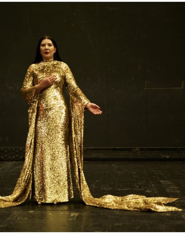 Woman in floor-length gold sequin gown with long sleeves stands in a dark space with her hand on her chest looking at camera.