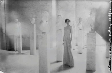 Model in long dress standing among classical white busts on pillars in a gallery by photographer Stefan Milev
