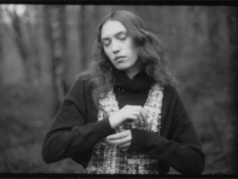 Model with long wavy hair wearing a dark turtleneck and patterned vest outdoors in a forest by Stefan Milev