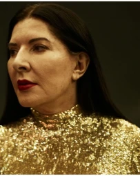 Portrait of Marina Abramovic with dark hair and red lipstick wearing a gold sequin top against a dark background.