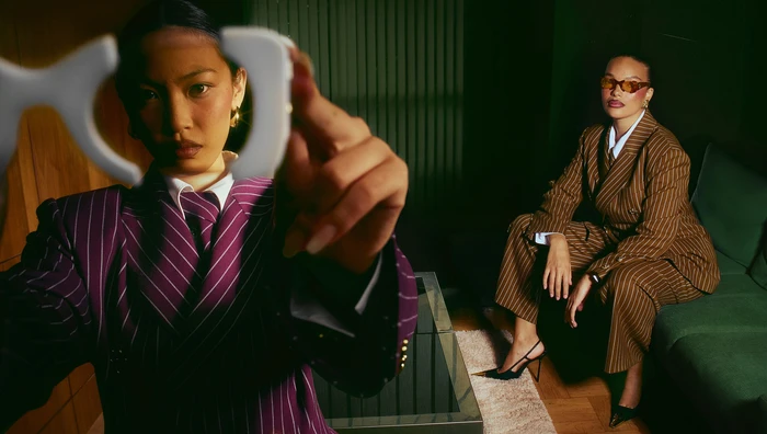Two women in tailored pinstripe suits holding up eyeglass frames in a dimly lit interior space in London by Bater and Street.