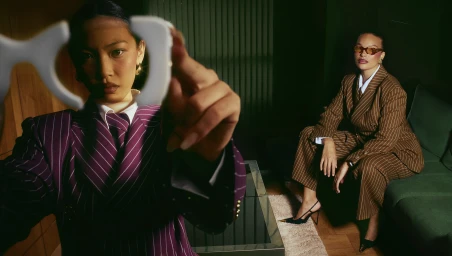 Two women in tailored pinstripe suits holding up eyeglass frames in a dimly lit interior space in London by Bater and Street.