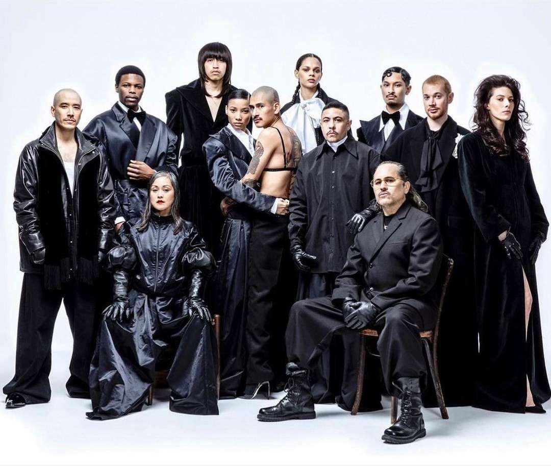 Group portrait of eleven fashionably dressed people in a photo studio, wearing black clothing and boots on a seamless white background.