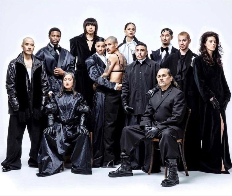 Group portrait of eleven fashionably dressed people in a photo studio, wearing black clothing and boots on a seamless white background.