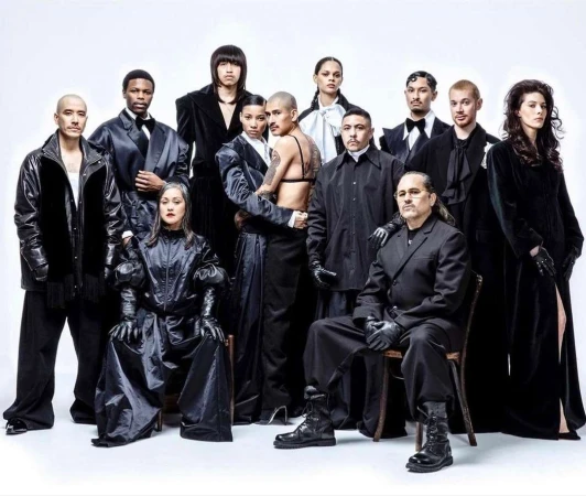 Group portrait of eleven fashionably dressed people in a photo studio, wearing black clothing and boots on a seamless white background.