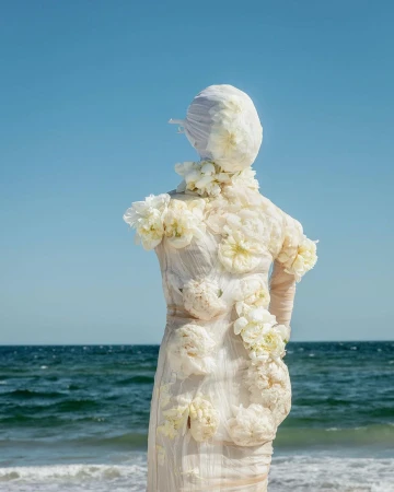 Figure covered in white floral appliqués and fabric standing against an ocean and clear sky background