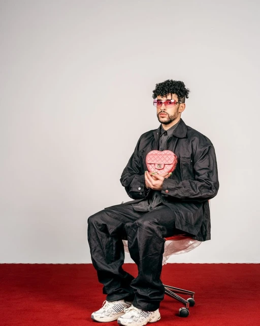 Bad Bunny sits on a red carpet wearing sunglasses and a black outfit holding a pink heart-shaped Chanel purse in a studio setting.