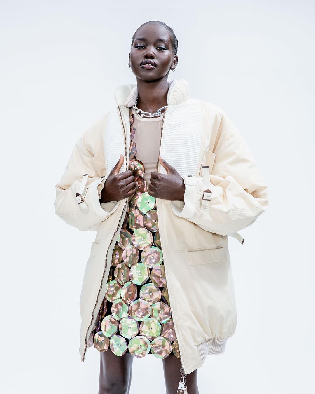 A Black female fashion model poses in a studio wearing a beige puffer coat, tan top, and a reflective disc mini skirt against a white backdrop.