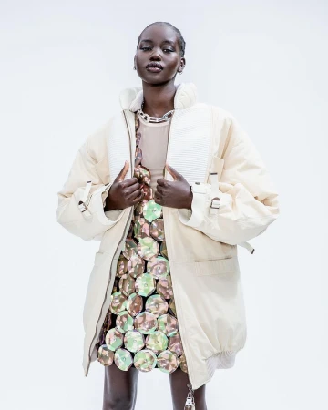 A Black female fashion model poses in a studio wearing a beige puffer coat, tan top, and a reflective disc mini skirt against a white backdrop.