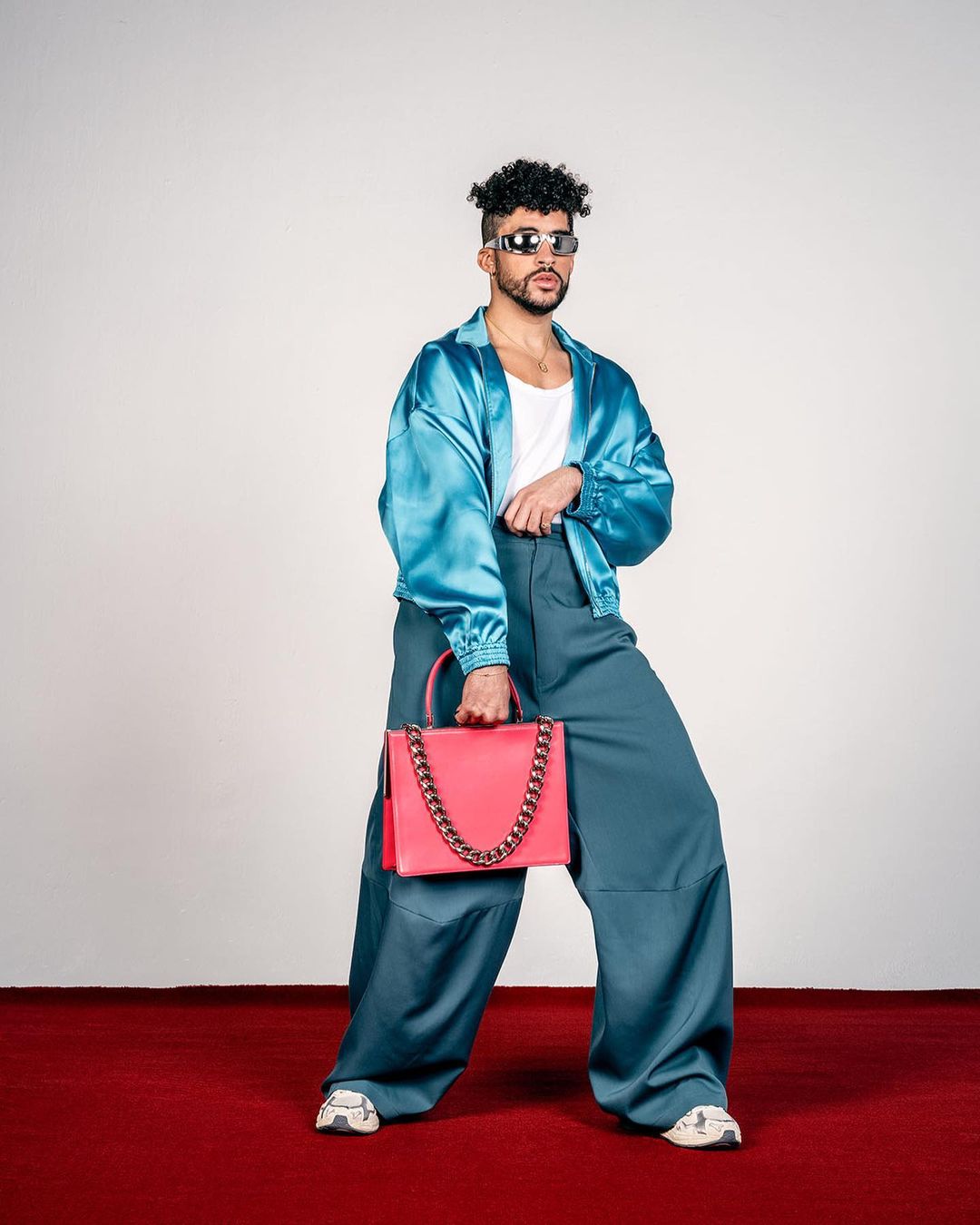 Bad Bunny strikes a pose in a studio, wearing a blue jacket and wide pants, holding a pink purse, and sporting black sunglasses.