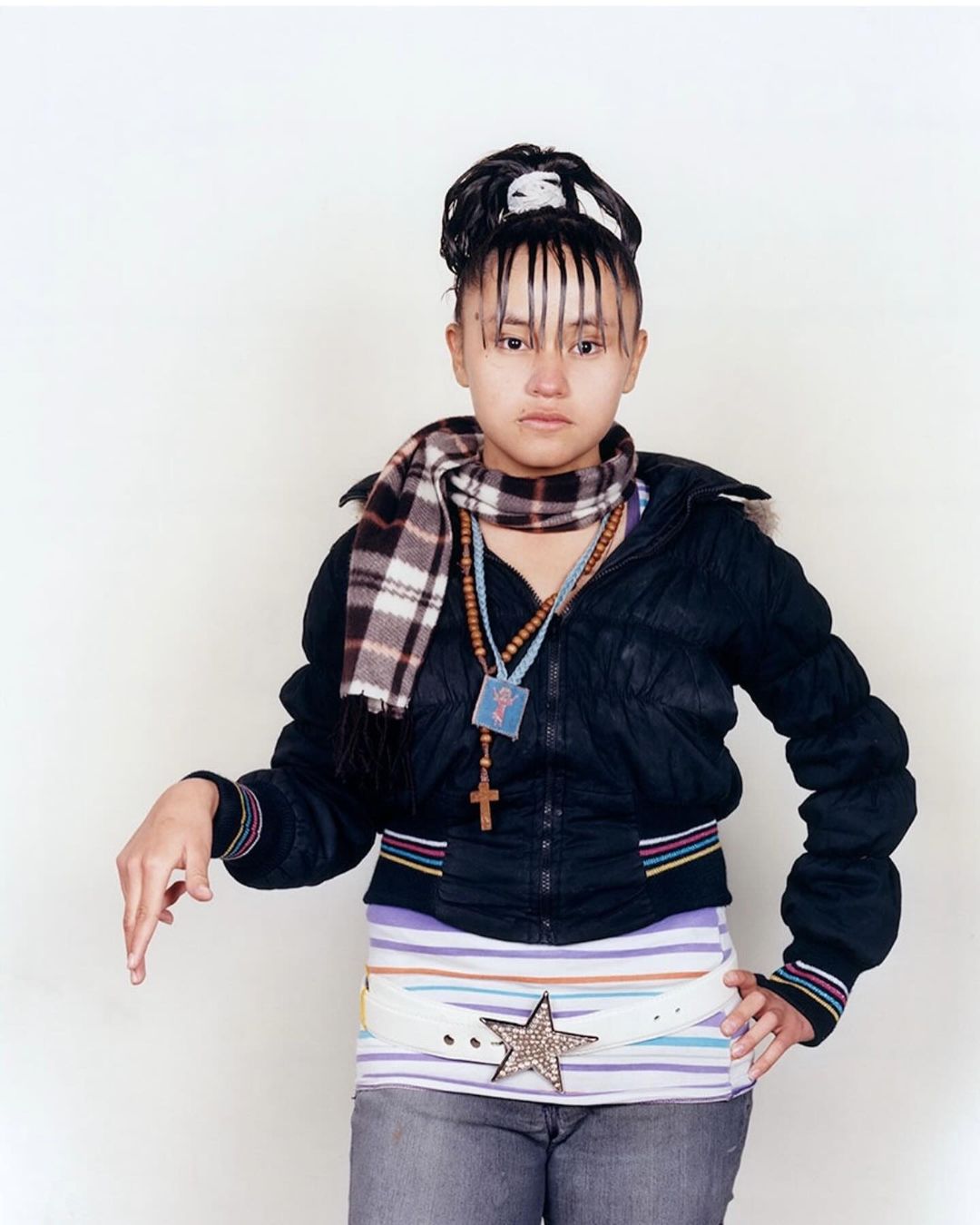 Teenage girl in a black jacket and grey jeans standing in front of a white wall. She is wearing a scarf, and a belt with a star.