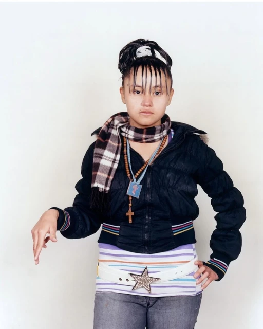 Teenage girl in a black jacket and grey jeans standing in front of a white wall. She is wearing a scarf, and a belt with a star.